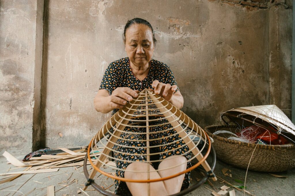 Elderly woman skillfully weaves a traditional conical hat indoors, showcasing cultural craftsmanship.
